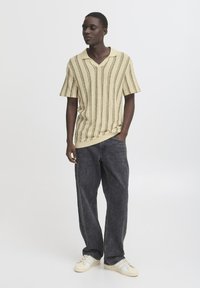 Strikket kortermet polo i lys gul med vertikale striper i mørkebrunt; kombinert med mørke jeans og kremfargede joggesko.