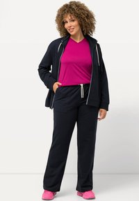 Marineblauwe trainingspak set met een ritssluiting jack, een roze v-hals shirt en bijpassende sweatpants met trekkoord. Heeft een zachte textuur en roze sneakers.