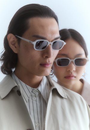 Naočale sa svijetlosivim okvirom i tamnim staklima, sa pravokutnim oblikom. Model nosi bež trenč kaput preko majice sa prugama.