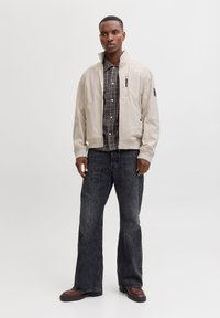 Veste bomber beige clair, poches zippées, chemise à carreaux noire en dessous, jean foncé et bottes marron. Texture lisse et coupe décontractée.