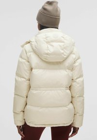 Lysfarvet pufferjakke med vandret quiltede sektioner, hætte og et logo på ryggen. Materialet fremstår glat og blankt.