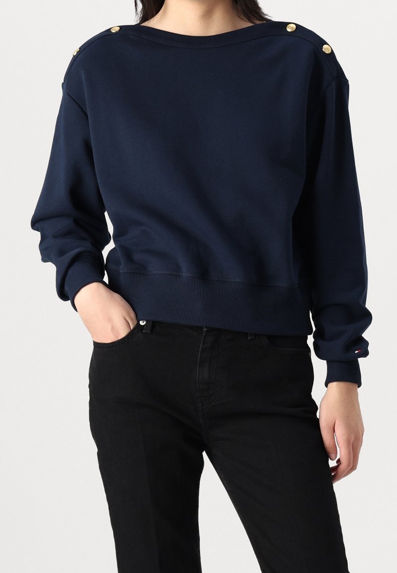 Femme portant un pull bleu marine à manches longues avec des boutons dorés aux épaules et un pantalon noir, main dans la poche, debout devant un fond blanc.