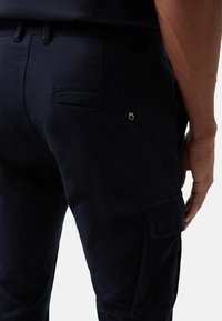 Achteraanzicht close-up van een persoon in een donkere marineblauwe broek met een achterzak en een zijcargozak, hand rustend naast de dij.