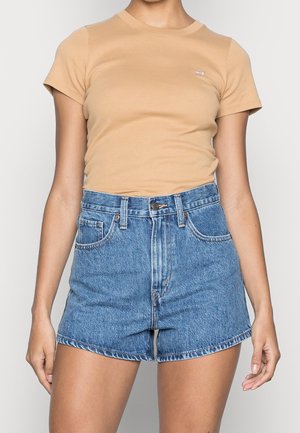 Femme portant un t-shirt beige à manches courtes rentré dans un short en denim bleu taille haute avec des poches devant et derrière.