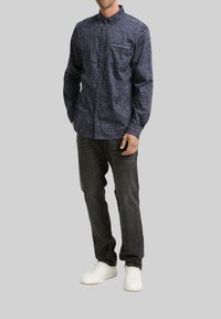 Chemise bleu marine à boutons avec un motif à pois multicolores, manches longues, poche poitrine avec bord blanc, portée avec un jean foncé et des baskets blanches.
