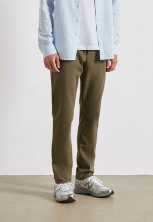 Pantalons en coton vert olive associés à une chemise boutonnée rayée bleu clair et des chaussures de sport grises sur un fond neutre.