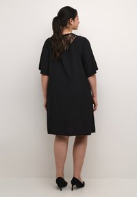 Vestido negro con mangas cortas y un detalle de encaje en la parte posterior. La tela parece suave con sutiles rayas verticales. Unos tacones completan el conjunto.