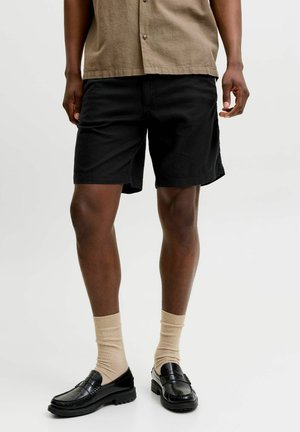 Mężczyzna ubrany w czarne casualowe szorty, beżowe skarpetki, czarne mokasyny i beżową koszulę z krótkim rękawem na białym tle.