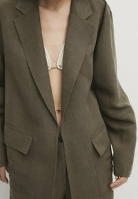 Olijfgroene linnen blazer met een notch-lapel, twee opgestikte zakken en opgerolde mouwen, gedragen over een crèmekleurige kanten bh, met een ontspannen pasvorm.