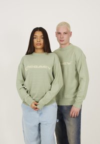 Grønne strikkede sweatre med en subtil tekstur og ribbede manchetter, der har ordet "LEGACY" i crema over brystet.
