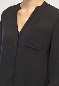 Black button-up shirt with a chest pocket and a narrow, rounded collar worn by a person with a visible neck and upper chest.