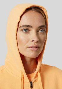 Bluza z kapturem w jasnym pomarańczowym kolorze, wykonana z miękkiego materiału. Wyposażona w zamek błyskawiczny i sznurek do regulacji, charakteryzuje się gładką teksturą i luźnym krojem.