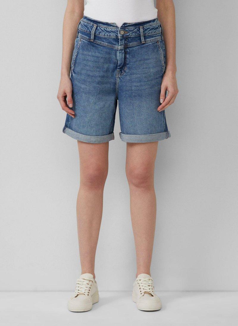 Shorts en jean bleu clair avec un ourlet roulé, dotés d'une fermeture à bouton à l'avant et de poches latérales, associés à un haut blanc et des chaussures claires.