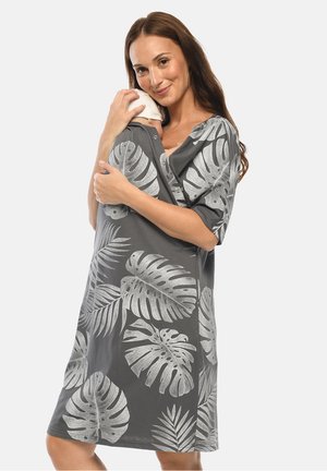 Woman wearing a gray dress with large white tropical leaf prints, holding and gently cradling a baby wrapped in a matching fabric.