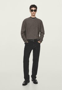 Pullover marrone a costine, jeans neri e scarpe nere. Gli accessori includono occhiali da sole scuri. Design semplice con una vestibilità casual e un contrasto di texture.