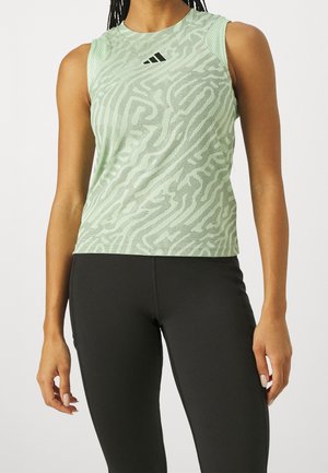 Žena nosící světle zelený sportovní top bez rukávů s vzorem a černé legíny, stojící s pažemi uvolněnými podél těla.