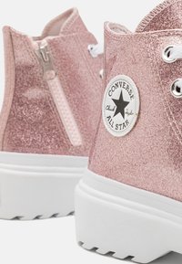 Różowe, błyszczące trampki Converse typu high-top z białą, grubą podeszwą, sznurówkami, zamkiem błyskawicznym po boku oraz naszywką logo "Converse All Star" na kostce.