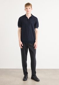 Polo shirt bleu marine avec des manches courtes légèrement bouffantes et des accents de rayures blanches sur les poignets. Accompagné d'un pantalon noir et de chaussures sombres.