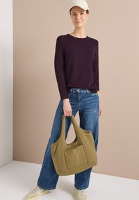 Olijfgroene, zachte nylon schoudertas met een geraffineerde bovenkant en een voertas, vastgehouden door een persoon in een donkerpaarse lange mouw shirt en blauwe spijkerbroek.