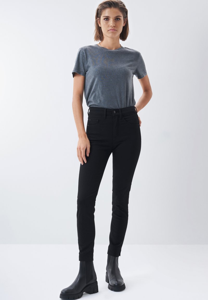 Černé skinny džíny s vysokým pasem, v kombinaci s přiléhavým šedým tričkem. Modelka má na sobě černé kotníkové boty. Jednoduchý design, minimální detaily.