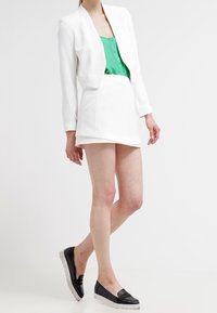 Bílý krátký sako přes zelenou bezrukošovou blůzu, doplněné bílou mini sukní a černými klouzavými botami s texturovanými akcenty.