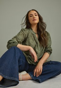 Olivengrünes Shirt mit floraler Stickerei, Knopfverschluss vorne und gerafften Ärmeln, kombiniert mit weit geschnittenen blauen Jeanshosen und beigen Sandalen.