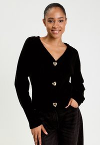 Schwarze Strickjacke mit tiefem V-Ausschnitt, drei goldenen herzförmigen Knöpfen und langen Ärmeln, aus weichem, strukturiertem Stoff gefertigt.