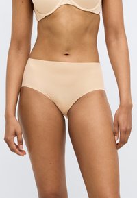 Γυναίκα που φοράει αόρατο μπεζ εσώρουχο υψηλής μέσης και αντίστοιχο σουτιέν, στέκεται μπροστά σε ένα απλό φωτεινό φόντο.