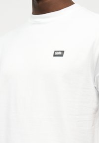 Κοντινό πλάνο ενός ατόμου που φοράει ένα απλό λευκό μπλουζάκι με στρογγυλή λαιμόκοψη και ένα μικρό μαύρο ορθογώνιο λογότυπο στο στήθος.