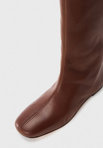 Botte en cuir marron, jusqu'au genou, avec un bout rond, une texture lisse, des détails de couture subtils et une silhouette élancée.