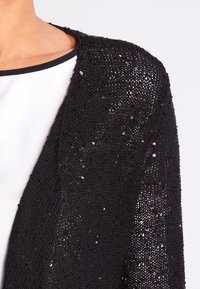 Černý pletený cardigan s detailním zdobením flitry, otevřeným předním designem a lehkou texturou, vrstvený přes bílé tričko.