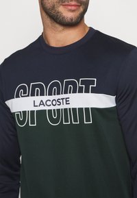 Camisa deportiva de manga larga en azul marino y verde con el texto "SPORT" en blanco en gran tamaño y una franja horizontal blanca con el logo de "LACOSTE". Textura de tejido suave.