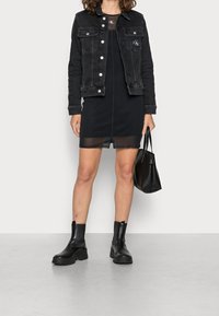 Svart denimjacka med silverknappar och CK-logotyp, kombinerad med en svart klänning med genomskinlig fåll, svarta ankelboots och en svart handväska.