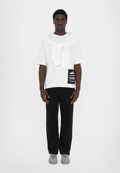 Hvid kortærmet t-shirt med en sort lap, parret med sorte bukser. En hvid sweater er draperet om halsen. Grå sko fuldender outfittet.