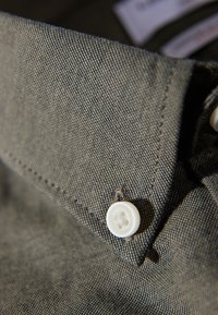 Collar de camisa caqui con una tela texturizada. Presenta un botón blanco redondo y un detalle de costura visible. Vista en primer plano centrada en los elementos de diseño.