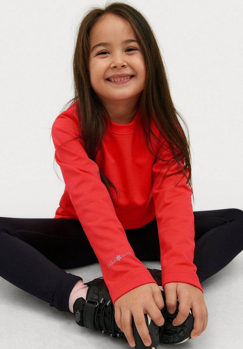 Rotes Langarmoberteil mit rundem Ausschnitt, kombiniert mit schwarzen Leggings und schwarzem Schuhwerk. Mit Logo-Detail am Ärmel.