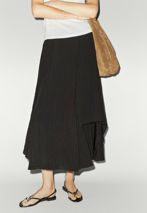 Persona che indossa una gonna asimmetrica nera, top bianco, sandali neri e porta una borsa a tracolla in camoscio marrone con le braccia incrociate.
