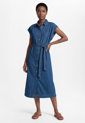 Kobieta w bezrękawną, zapinaną na guziki sukienkę midi z denimu z przewiązanym paskiem w talii i brązowymi sandałami typu slide, stoi z jedną ręką na biodrze.