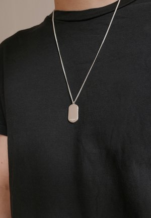 Silver chain necklace with a rectangular dog tag pendant, featuring a smooth surface and rounded edges, worn over a black t-shirt.