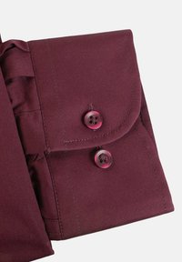 Telaje marrón con textura suave, con un cierre de solapa abotonado y dos botones a juego. Líneas limpias y diseño ajustado.