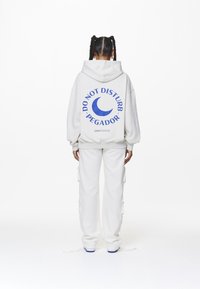 Sudadera blanca con el texto azul "NO MOLESTAR PEGADOR" y un gráfico de luna creciente en la parte posterior, combinada con pantalones blancos a juego.