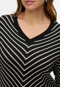 Pull rayé noir et crème avec un col en V, présentant un motif en chevron et un design de tricot texturé. Accentué avec des côtes noires.