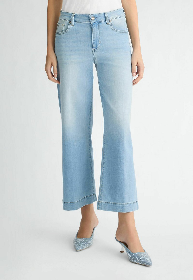 Jean large bleu clair en denim, avec une taille haute, un ourlet effiloché et un design à cinq poches. Associé à des mules à talons embellies.