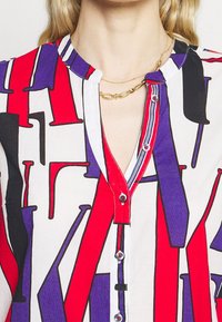 Knapp-up skjorta med ett färgstarkt abstrakt mönster i röd, svart och lila på en vit bakgrund; har en enkel krage och synliga knappar.