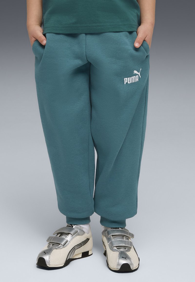 Enfant portant un pantalon de survêtement Puma bleu-vert et des baskets Puma argentées avec des bandes Velcro, debout les mains dans les poches.