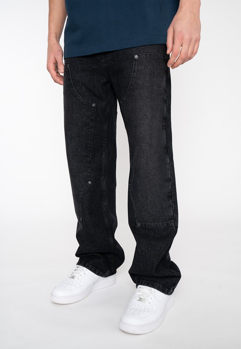 Schwarze Jeans aus Denim mit lockerer Passform, verziert mit gebogenen Nähten und silbernen Hardware-Details. Kombiniert mit weißen Sneakers.