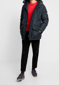 Muž v černé prošívané bundě přes červený svetr, černých kalhotách, bordó ponožkách a tmavě modrých semišových botách stojící proti jednobarevnému pozadí.