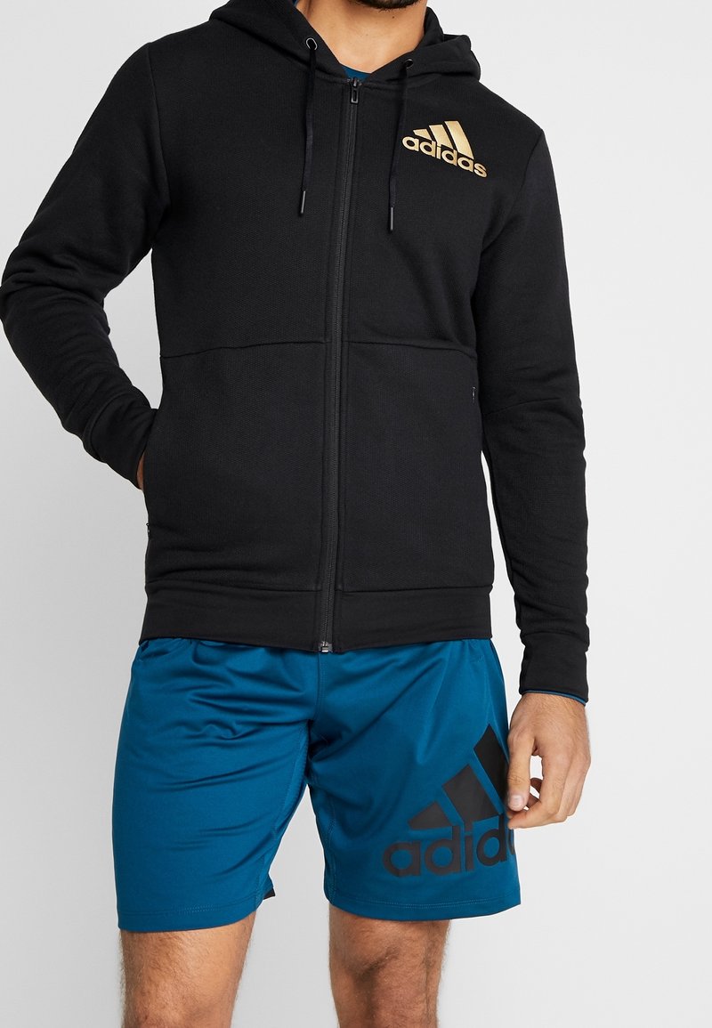 Sudadera negra con cremallera y logo de adidas dorado, combinada con pantalones cortos deportivos azules que tienen un gráfico negro de adidas en la pierna. Textura de tela suave.
