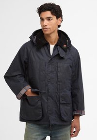 Casaco encerado azul-marinho com colarinho de veludo cotelê, fecho frontal com botões, dois bolsos grandes e forro xadrez visível nas mangas.