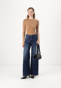 Top à manches longues beige avec des détails froncés, accompagné d'un jean large bleu foncé. Tient un petit sac à main noir et porte des talons noirs.